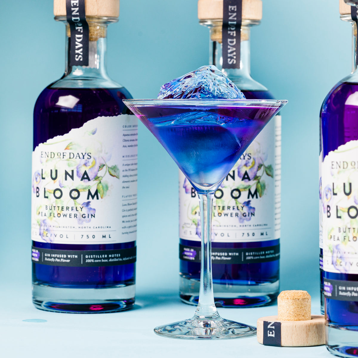 Blue and purple Luna Bloom gin martini with several bottles on a light blue background