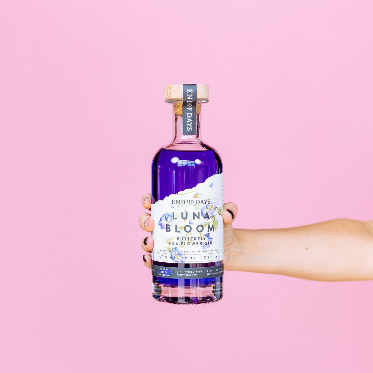 Hand holding a bottle of Luna Bloom purple gin against a soft pink background