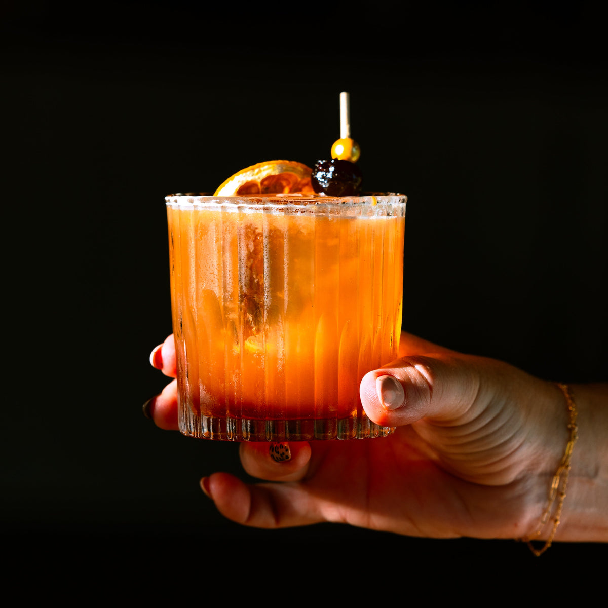 A hand holding a frosted glass tumbler filled with an orange cocktail, garnished with a citrus slice and a dark olives on a pick, against a solid black background.