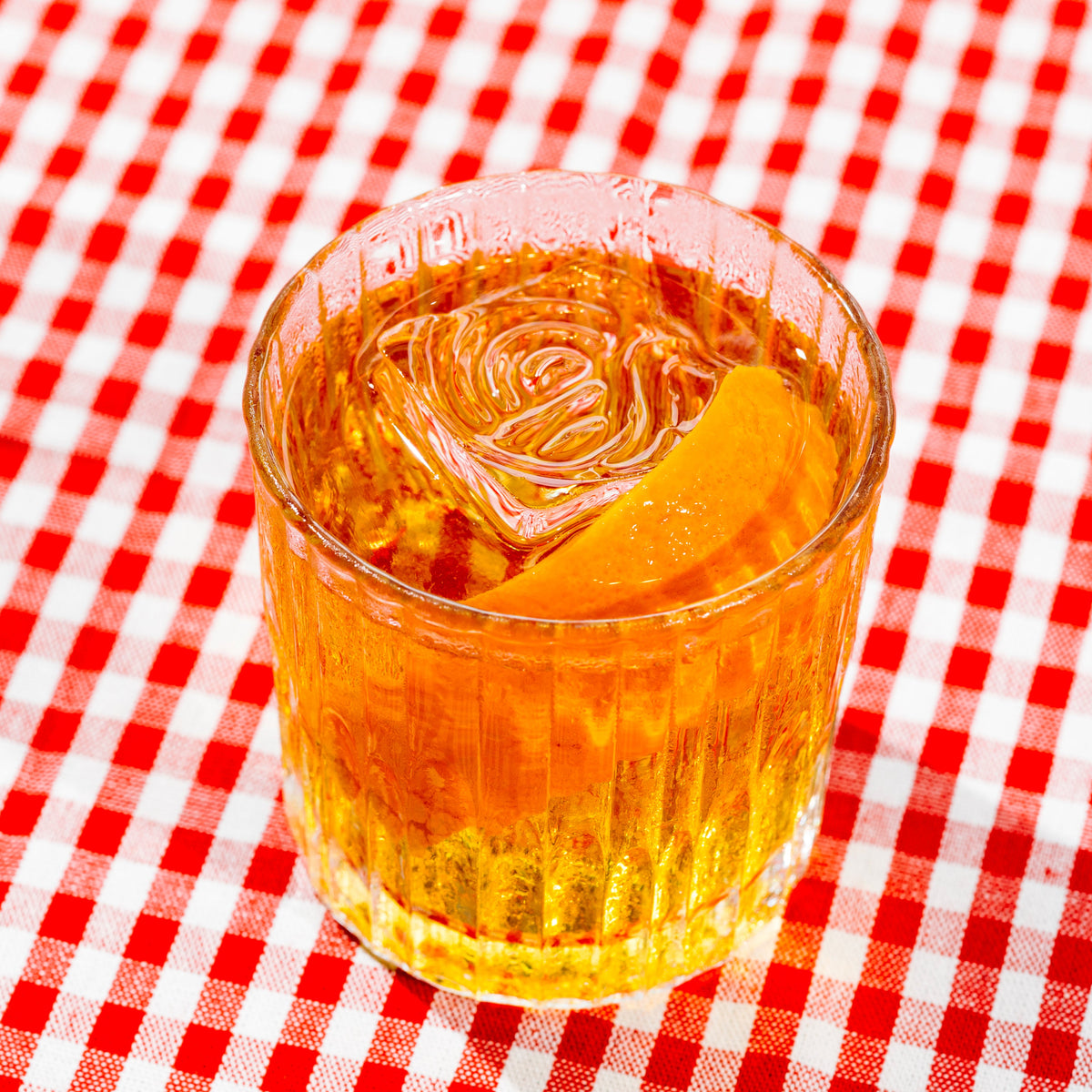 A clear glass tumbler filled with amber-orange liquid and a large rose-shaped ice cube, set on a red-and-white checkered tablecloth.
