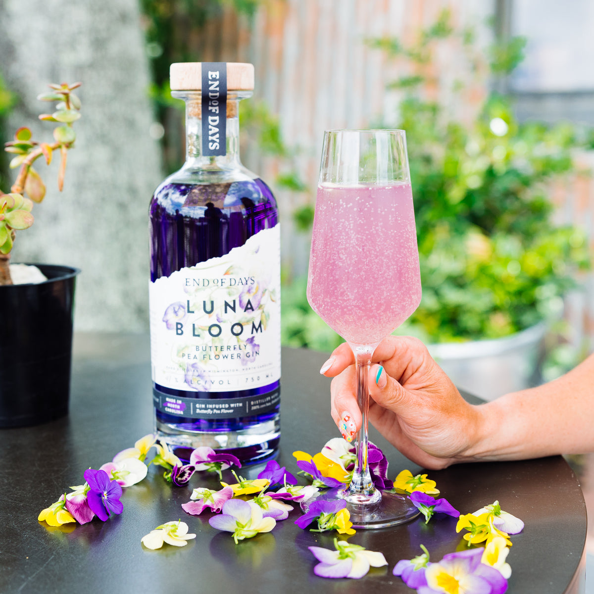 Pink color-changing gin cocktail made with Luna Bloom, surrounded by colorful flowers outdoors
