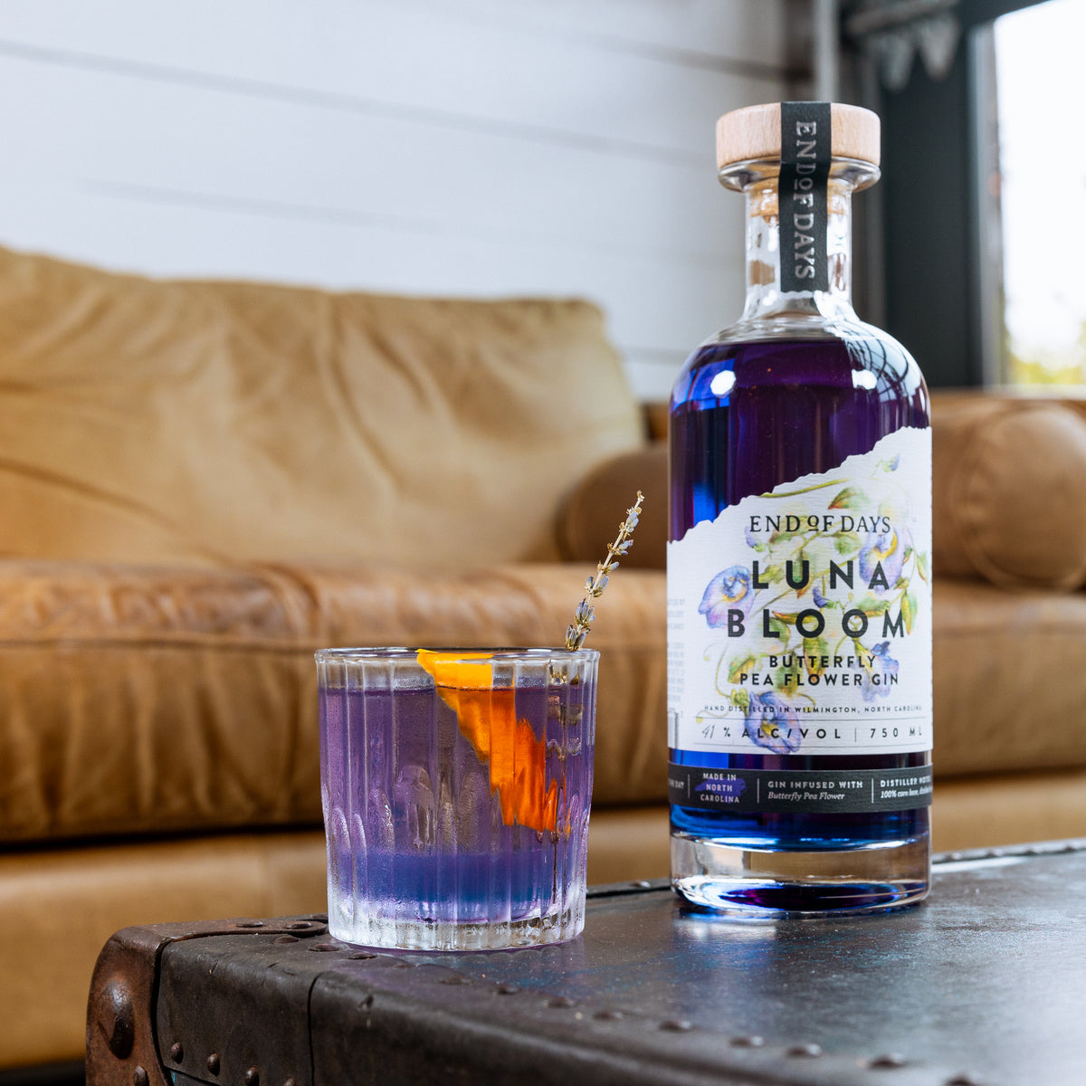 Luna Bloom gin bottle beside a purple gin cocktail with an orange garnish in a cozy lounge