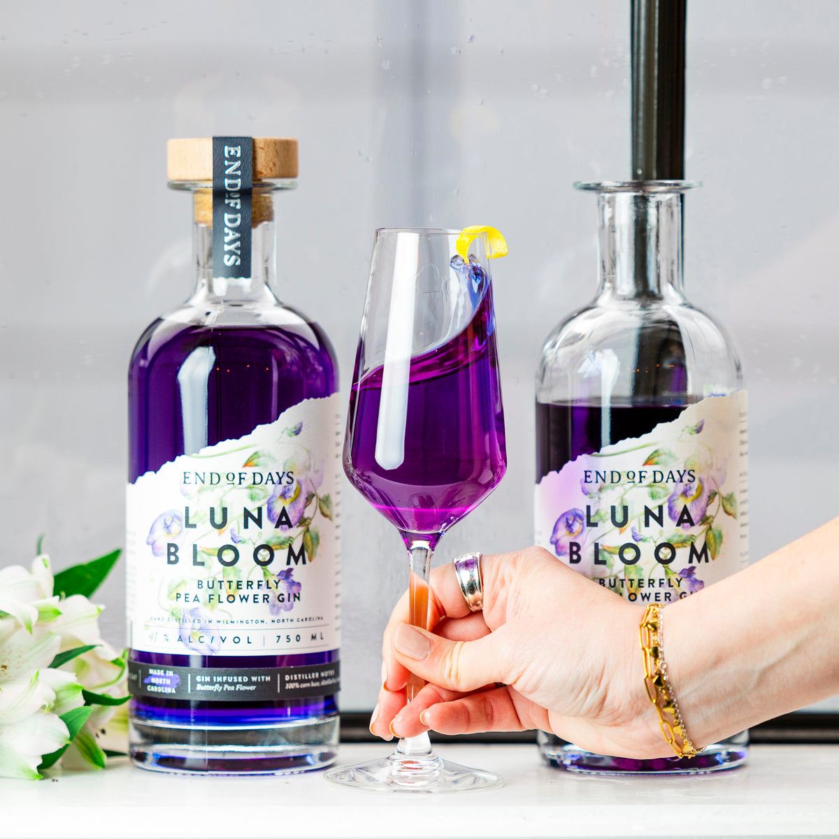 Two bottles of Luna Bloom purple gin with a poured glass and white flowers in a bright cocktail setting