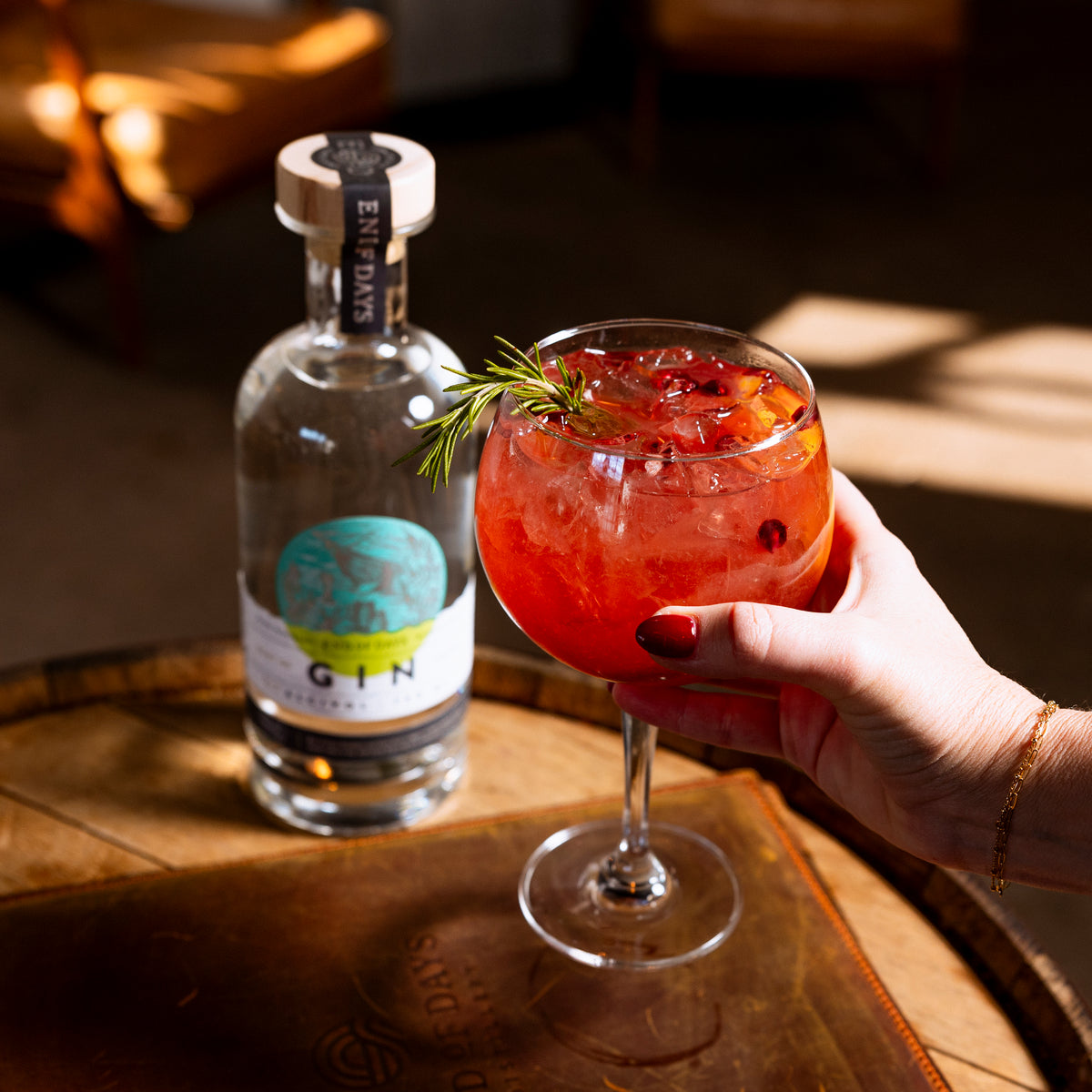 Hand holding a red gin cocktail garnished with rosemary beside a bottle of Port of Entry Gin