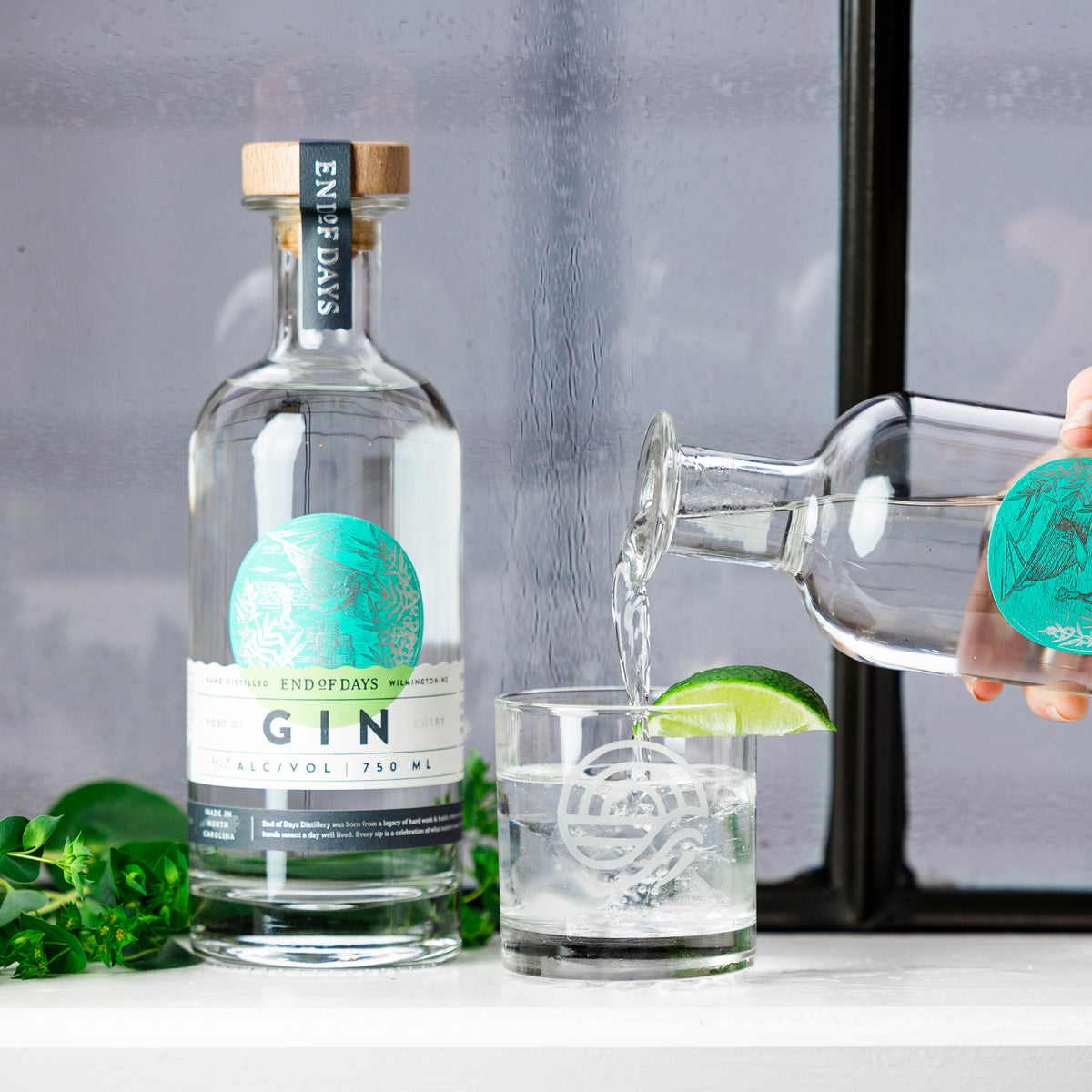 Port of Entry Gin poured over ice with a lime wedge, beside the bottle and fresh herbs